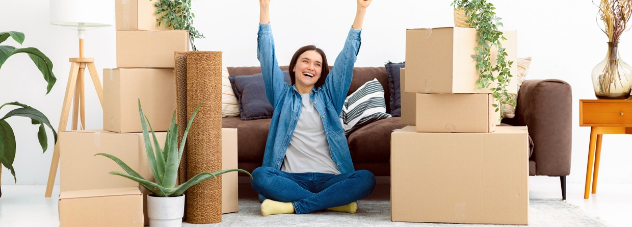 Vrolijke jonge vrouw zit blij op de grond tussen haar verhuisdozen in haar nieuwe huis
